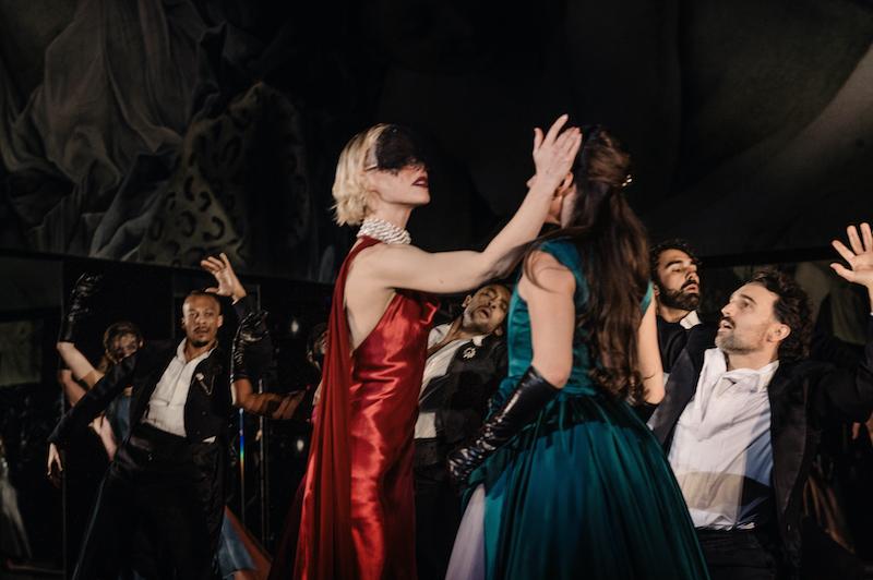 A masked woman approaches another, while surrounded by male dancers